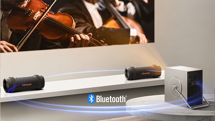 LG CineBeam Q projector is projecting concert scene onto the wall and two units of xboom Grab are placed on both side below the screen.